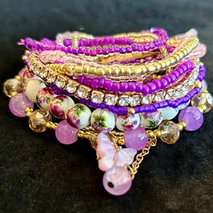 May include: A stack of beaded bracelets in shades of purple, pink, and gold. The bracelets feature various bead types, including small seed beads, larger round beads, and crystal accents. A charm with a pink butterfly pendant hangs from the stack.