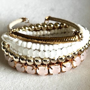 May include: A stack of five bracelets featuring a mix of gold-tone metal beads, smooth white spherical beads, translucent white beads, and a row of pale pink faceted stones set in gold-tone metal frames, finished with a textured gold-tone bar.