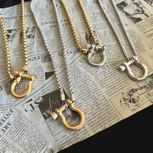 May include: Four necklaces with a gold or silver finish are displayed on a newspaper background. Each necklace features a chain and a U-shaped pendant with a screw detail. The necklaces are a mix of gold and silver tones.