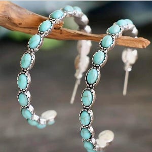 May include: A pair of silver-toned hoop earrings. Each earring features a series of oval, turquoise-colored stones set in a decorative frame. The earrings are displayed against a blurred background, highlighting their design.