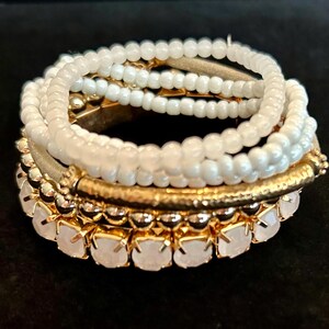May include: A stack of bracelets featuring a variety of designs. The bracelets include white beaded strands, gold-toned metal bands, and a bracelet with white, faceted stones. The bracelets are stacked together, creating a layered look.