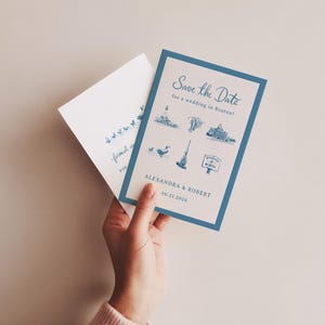 K&ouml;nnte beinhalten: Eine blau-wei&szlig;e Hochzeitseinladung mit dem Text "Save the Date" und Illustrationen von Wahrzeichen von Boston. Die Einladung enth&auml;lt die Namen "Alexandra & Robert" und das Datum "06.21.2026". Ein passender Umschlag ist ebenfalls zu sehen.