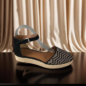 May include: Black and beige espadrille wedge sandals with a chevron pattern on the toe. The sandals have a black ankle strap with a silver buckle and a woven jute sole. The brand name is visible on the insole.