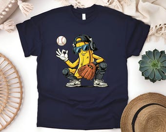 May include: Navy blue t-shirt featuring a cartoon banana catcher wearing sunglasses, headphones, and a baseball glove. The banana is squatting, holding a baseball, and making an 'okay' hand gesture. A baseball is floating in the air.