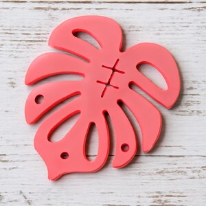 Puede incluir: Un posavasos en forma de hoja de color coral con un diseño cosido en el centro. El posavasos tiene varios agujeros y una estética tropical única. Está hecho de un material sólido y descansa sobre una superficie de madera blanca.