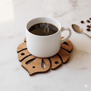 Puede incluir: Una taza de cerámica blanca llena de café oscuro se encuentra sobre un posavasos de madera con forma de hoja de monstera. El vapor se eleva del café. Una cuchara plateada y granos de café están cerca.