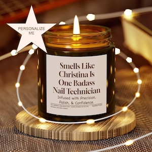 May include: A lit candle in a dark glass jar with a gold rim, on a wooden coaster. The label says "Smells Like Christina Is One Badass Nail Technician." Fairy lights encircle the candle, creating a warm glow.
