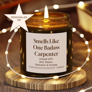 Può includere: Una candela accesa in un barattolo di vetro ambrato con un'etichetta bianca che riporta il testo: Smells Like One Badass Carpenter, Infused With Skill, Passion, Dedication & Purpose. Eco-Friendly And Non-Toxic, Hand-Poured In The USA, Smokin Wicks.