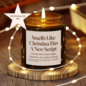 May include: A lit candle in a dark green glass jar with a label that reads "Smells Like Christina Has A New Script." The candle sits on a wooden coaster, surrounded by string lights. The text "Personalize Me" is on a star.