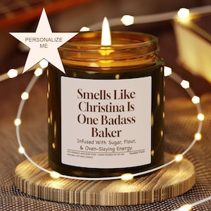 Könnte beinhalten: Eine brennende Sojawachskerze in einem Glas mit einem Etikett mit der Aufschrift "Smells Like Christina Is One Badass Baker". Die Kerze steht auf einem Holzuntersetzer und ist von Lichterketten umgeben.
