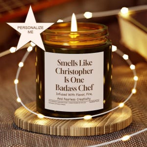 May include: A lit candle in a glass jar on a wooden coaster. The candle has a label that reads "Smells Like Christopher Is One Badass Chef." A string of lights and a star-shaped graphic with "Personalize Me" are also in the image.