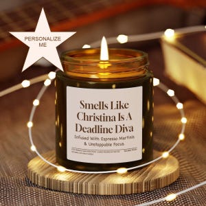 Peut inclure: Une bougie allumée dans un pot vert foncé avec une étiquette blanche qui dit "Smells Like Christina Is A Deadline Diva". La bougie est posée sur un dessous de verre en bois, entourée de guirlandes lumineuses. Un signe en forme d'étoile dit "Personalize Me".