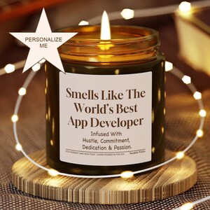 May include: A lit candle in a glass jar with the text "Smells Like The World's Best App Developer." The candle is on a wooden coaster and surrounded by string lights. The label also says "Infused With Hustle, Commitment, Dedication & Passion."