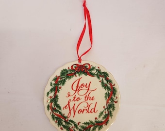 May include: Circular Christmas ornament with a cream background and a red hanging ribbon. It displays a green wreath with red ribbons and the words "Joy to the World" in decorative script.