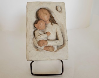 May include: A cream-coloured relief sculpture of a mother and child. The figures are carved with simplified features and painted in neutral tones. The sculpture is mounted on a dark metal stand.