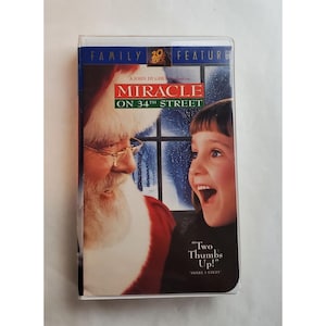 May include: A VHS tape for the movie "Miracle on 34th Street." The cover features Santa Claus and a young girl with the movie title in red and green. The text "Two Thumbs Up!" is also visible.