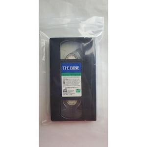 Può includere: Una videocassetta VHS nera, sigillata in un sacchetto di plastica trasparente, con il titolo "THE BIBLE" su un'etichetta blu e bianca. Il nastro ha dimensioni e forma standard, comunemente utilizzato per le registrazioni video.