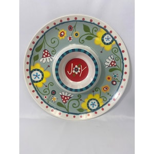 以下が含まれることがあります： 中央にディップボウルが付いた丸いセラミック製のサービング皿。皿は、黄色、赤、緑の花柄とつる柄の淡いティール色の背景が特徴です。ディップボウルは赤色で、白い文字で「JOY」と書かれています。