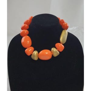 Op de afbeelding: Een opvallende ketting met grote, afgeronde kralen in helder oranje en goud. De ketting wordt getoond op een zwarte mannequin, wat het gedurfde kleurcontrast en het unieke ontwerp benadrukt. Een perfect accessoire.