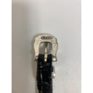 Op de afbeelding: Close-up van een zilverkleurige gesp met het jaar "&copy;1994" erop gestempeld. De gesp is bevestigd aan een zwarte, getextureerde riem.