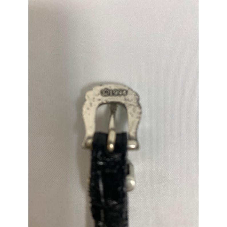 Op de afbeelding: Close-up van een zilverkleurige gesp met het jaar "&copy;1994" erop gestempeld. De gesp is bevestigd aan een zwarte, getextureerde riem.