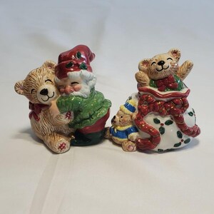 May include: Two ceramic Christmas figurines. One features Santa Claus hugging a teddy bear, both with smiling faces. The other is a Christmas sack with a teddy bear popping out. The sack is decorated with red ribbon and holly.