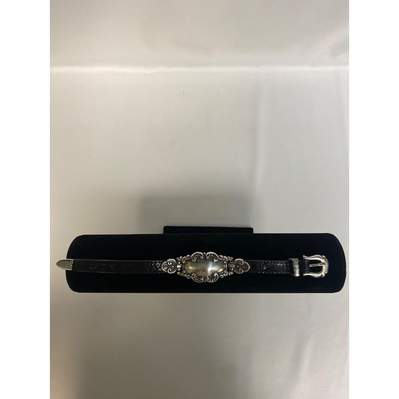 Op de afbeelding: Een zwart leren armband met een zilverkleurig middelpunt en een gesp. Het middelpunt heeft een ovale vorm met sierlijke details. De armband wordt gepresenteerd op een zwart fluwelen oppervlak.