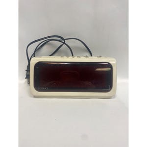May include: Cream-colored Cosmo digital alarm clock with a red display. The clock has a rectangular shape with rounded corners and several buttons on top. The power cord is black.