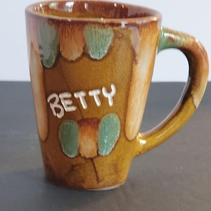 Könnte beinhalten: Eine handgefertigte Keramiktasse mit brauner und gelber Glasur. Die Tasse trägt den Namen "BETTY" in weißen Buchstaben und dekorativen grünen und braunen Akzenten. Der Griff hat eine passende gelbe Farbe.