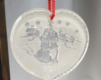 May include: Clear heart-shaped ornament with a winter scene and the year 2001 etched on the surface. The ornament is suspended by a red cord. The winter scene depicts a couple, a house, trees, and snowflakes.