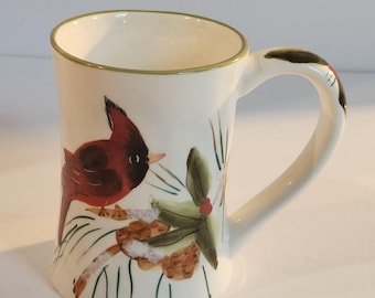 May include: A white ceramic mug with a red cardinal bird design. The mug features a green rim and handle accents, with a painted illustration of a bird perched on a branch with leaves and pinecones.