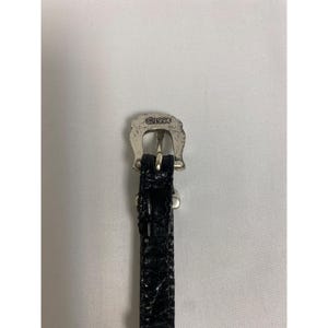 Op de afbeelding: Close-up van een zwarte leren riem met een zilverkleurige gesp. De gesp heeft de vorm van een hoefijzer en heeft de tekst "&copy;1994" gegraveerd. De riem heeft een textuur met krokodillenpatroon.