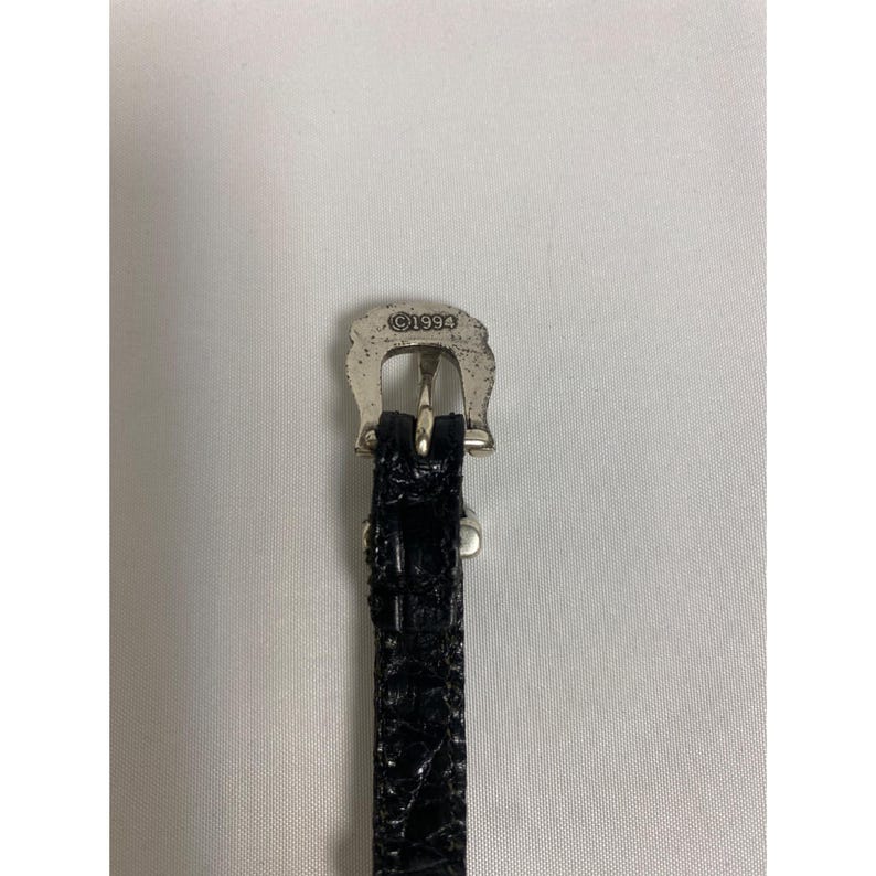 Op de afbeelding: Close-up van een zwarte leren riem met een zilverkleurige gesp. De gesp heeft de vorm van een hoefijzer en heeft de tekst "&copy;1994" gegraveerd. De riem heeft een textuur met krokodillenpatroon.