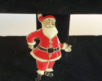 May include: A festive Santa Claus brooch in red, white, black, and gold. The brooch depicts a smiling Santa with a white beard, red suit, and black belt. The brooch is made of metal and has a pin fastening on the reverse.