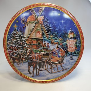 May include: Round tin with a Christmas scene featuring a windmill, horse-drawn sleigh, and a sign that reads "Jacobsen's Bakery Butter Cookies." The tin has a red and gold border and a winter landscape design.
