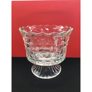 Puede incluir: Un cuenco de vidrio transparente con un borde festoneado y un patrón geométrico texturizado. El cuenco se asienta sobre una base de pedestal corta y acanalada. El fondo es rojo liso.