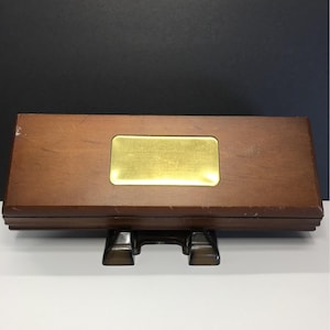 May include: A rectangular wooden box with a brass-colored metal plate in the center. The box is brown and rests on a black stand. The background is a dark color.