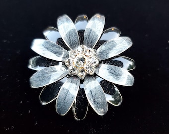 May include: A silver-toned flower brooch with a central cluster of clear rhinestones. The brooch features layered petals with a two-tone design of silver and black, set against a black background. A decorative accessory.