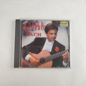 May include: CD case featuring Angel Romero playing a classical guitar. The album cover has the text "ANGEL ROMERO PLAYS BACH" in red and white. The image shows Romero in a tuxedo, with a red flower on his lapel.