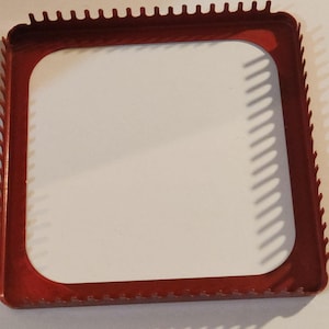 May include: A red, square-shaped metal frame with a white interior. The frame has a series of small, evenly spaced teeth along its outer edges. The corners of the frame are rounded.