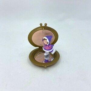 Peut inclure: Ornement de Noël vintage représentant un enfant patinant à l'intérieur d'un compact doré. L'enfant porte un bonnet violet et une veste rose. Le compact est ouvert, révélant un miroir et un intérieur rose.
