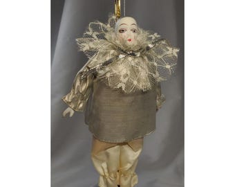 May include: A vintage clown doll ornament with a porcelain face, wearing a silver and cream-coloured outfit. The doll has a lace collar and is suspended by a gold string, ideal for festive decorations.