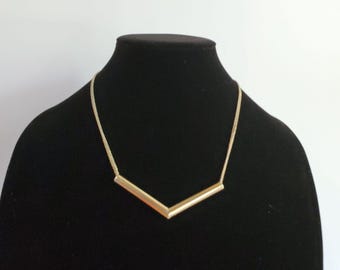 May include: A gold-coloured necklace with a V-shaped pendant. The pendant is made of two straight bars joined at an angle. The necklace is displayed on a black mannequin against a white background.