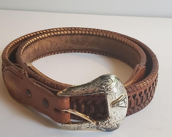 May include: A brown leather belt with a silver-colored buckle. The belt has a woven design along the edges and a smooth leather interior. The buckle features an engraved pattern and a silver finish.