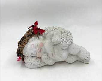 May include: A sleeping cherub figurine, crafted from light gray stone. The cherub is adorned with a small, brown wreath and a red flower. The cherub's cheeks are painted pink, and it has a textured wing.