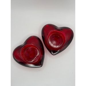 May include: Two heart-shaped, ruby red glass candle holders. Each holder has a circular indentation to hold a small candle. The glass is smooth and glossy, with a deep red colour. The candle holders are a romantic decorative item.