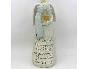 May include: A whimsical figurine of a bunny angel, holding a golden star. The bunny wears a light blue scarf and has white wings. The figurine has the text "He telleth the number of the Stars; He calleth them all by their names."