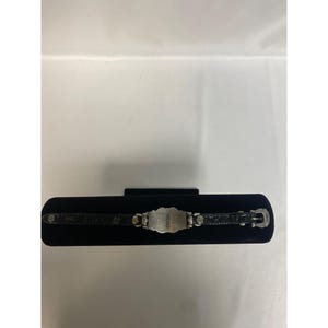 Op de afbeelding: Een zilverkleurige armband met een rechthoekig middengedeelte en een zwarte leren band. Het middengedeelte heeft een sierlijk ontwerp en het woord "Guess" is zichtbaar. De armband wordt gepresenteerd op een zwart fluwelen oppervlak.