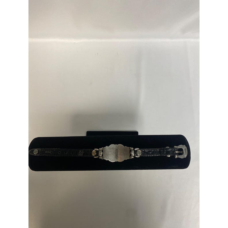 Op de afbeelding: Een zilverkleurige armband met een rechthoekig middengedeelte en een zwarte leren band. Het middengedeelte heeft een sierlijk ontwerp en het woord "Guess" is zichtbaar. De armband wordt gepresenteerd op een zwart fluwelen oppervlak.