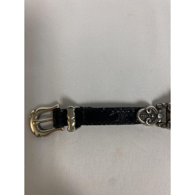 Op de afbeelding: Een zwarte leren riem met een zilverkleurige gesp en decoratieve elementen. De riem heeft een getextureerd oppervlak en een klassiek ontwerp. De gesp heeft een eenvoudige, gebogen vorm.